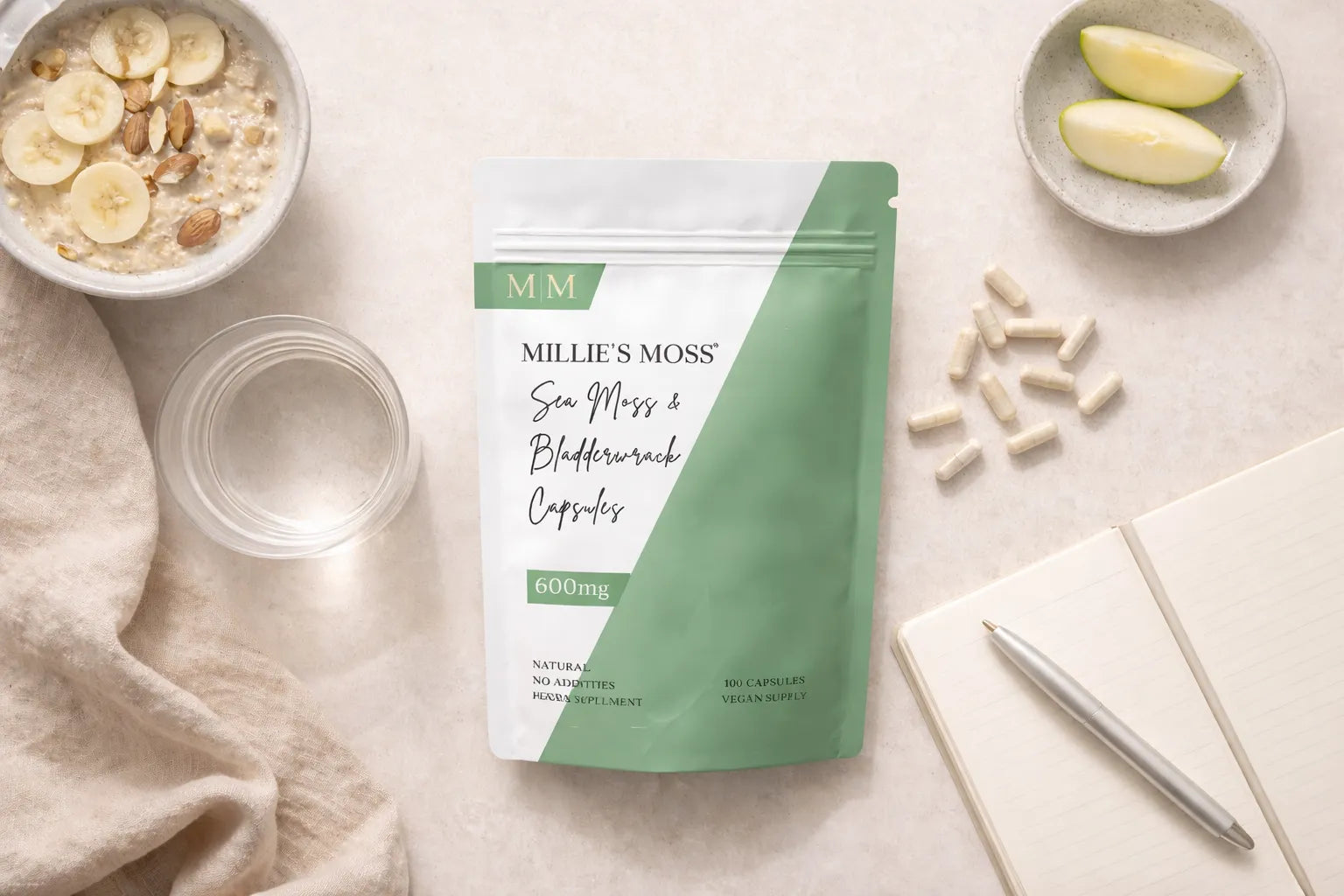 Flat lay of sea moss and bladderwrack capsules with breakfast bowl, apple slices, and notebook for a daily wellness routine.